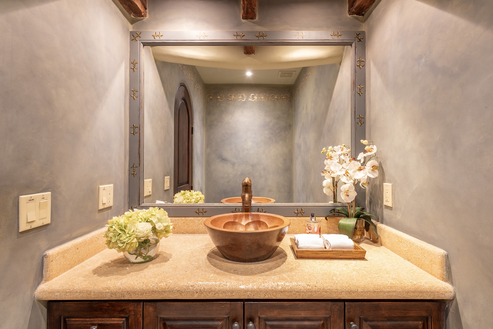 Refresh in your elegantly appointed bathroom with a unique copper vessel sink, hand-polished vanity, and rustic wood beam ceiling framed by soft lighting.