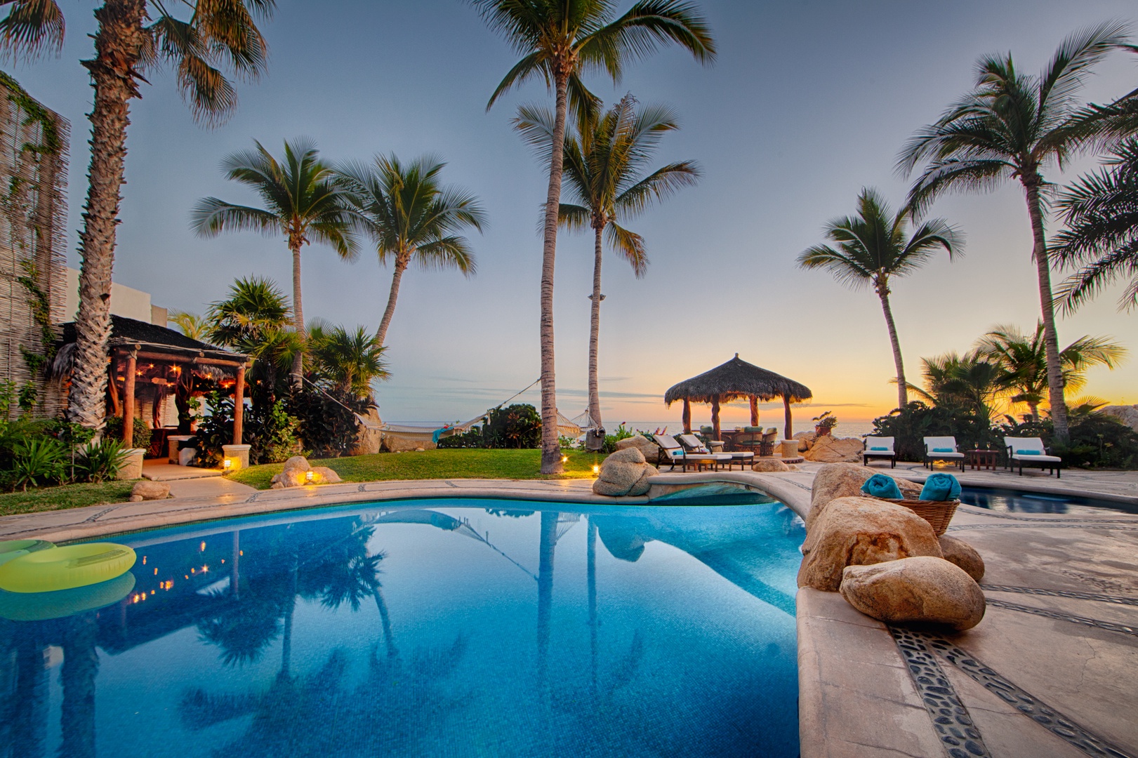 Tropical paradise pool area with palm trees, thatched gazebo, and stunning sunset views creating the perfect beachside retreat.