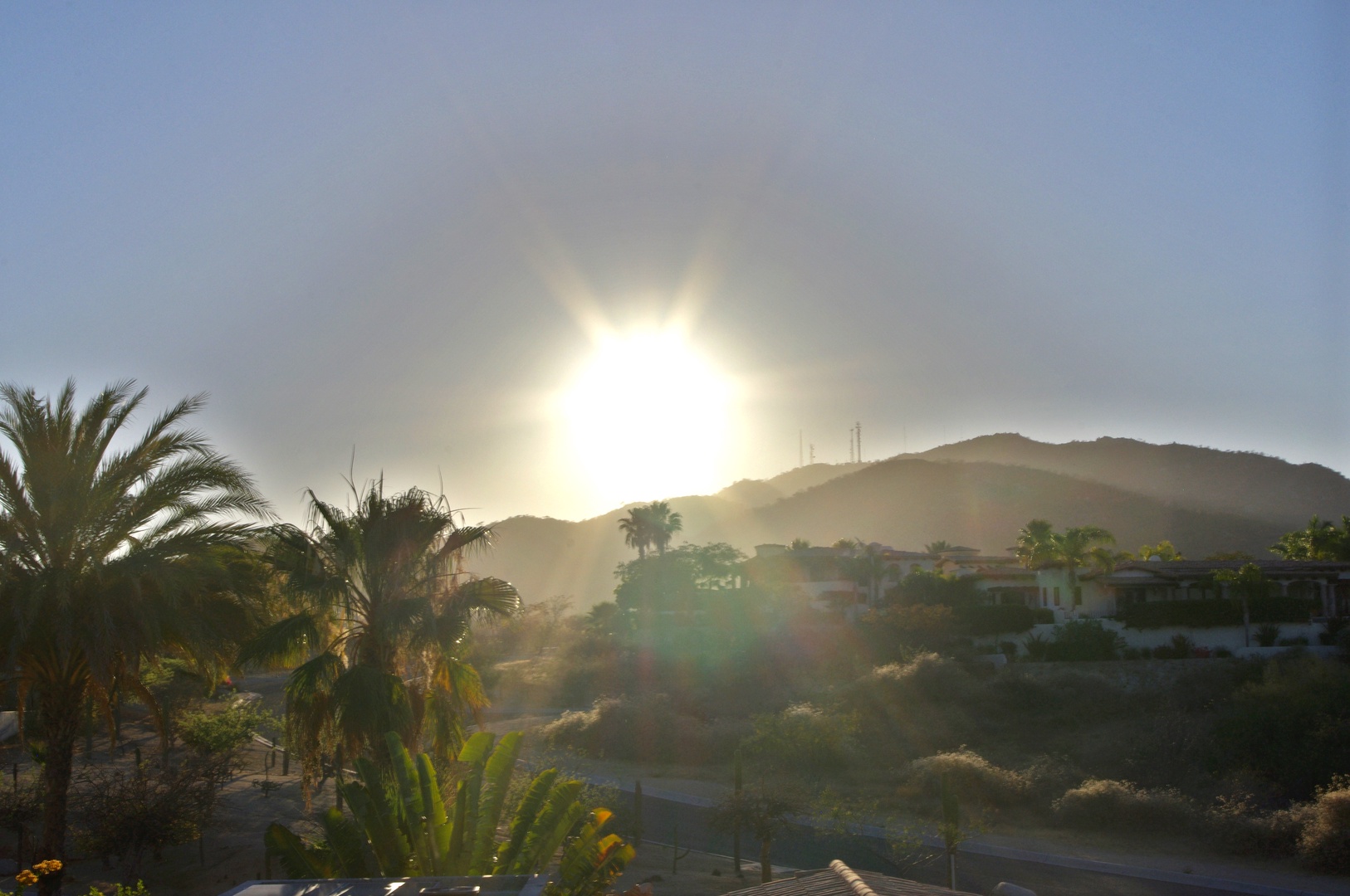 Sunrise illuminates the hillside neighborhood and palm-dotted landscape in the surrounding area, with the sun breaking over distant mountains in soft morning light.