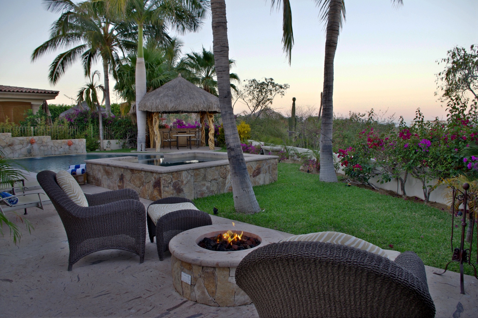 Sunset illuminates the landscaped garden with tropical palms, vibrant flowering plants, and a thatched palapa gazebo overlooking a stone hot tub and fire pit—a tranquil retreat within the property grounds.