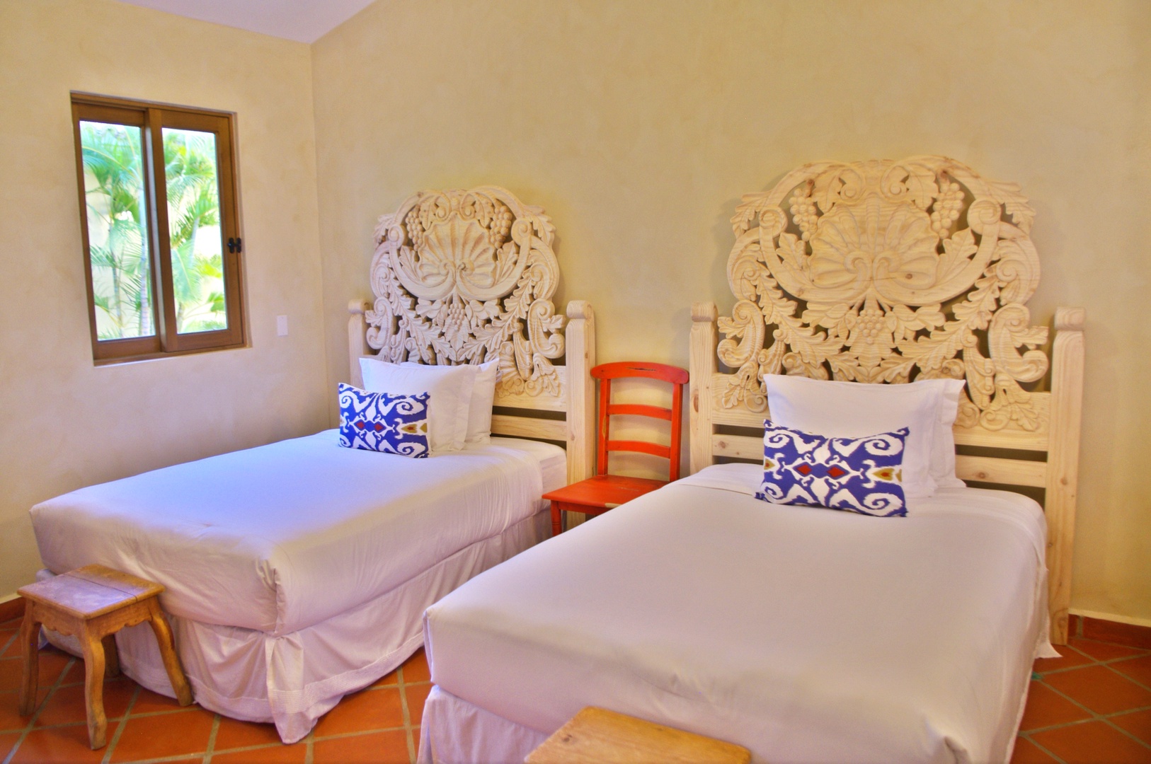 Settle into twin beds topped with artisan-carved headboards and vibrant accent pillows. Natural light streams in, complementing the warm tile floors and bright, airy walls.
