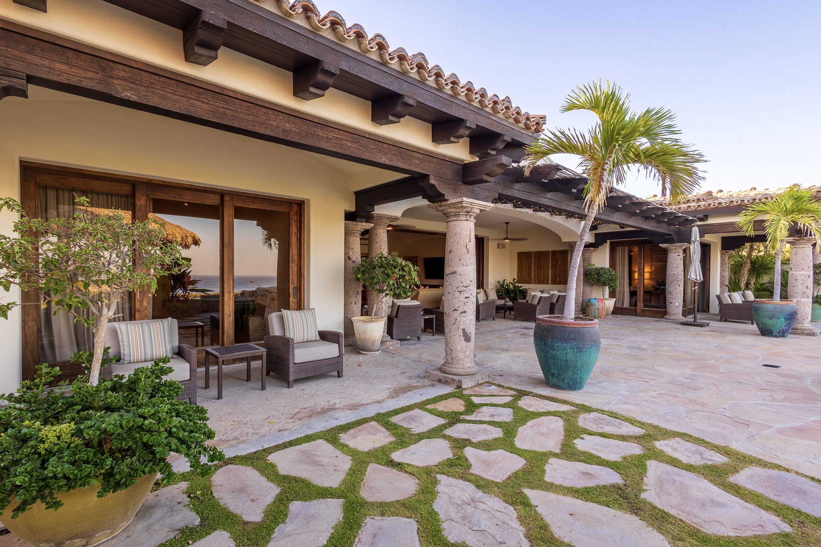 Elegant Spanish-colonial architecture with terracotta tiles and rustic wooden beams frames an inviting patio featuring comfortable seating, lush tropical palms, and decorative turquoise planters.