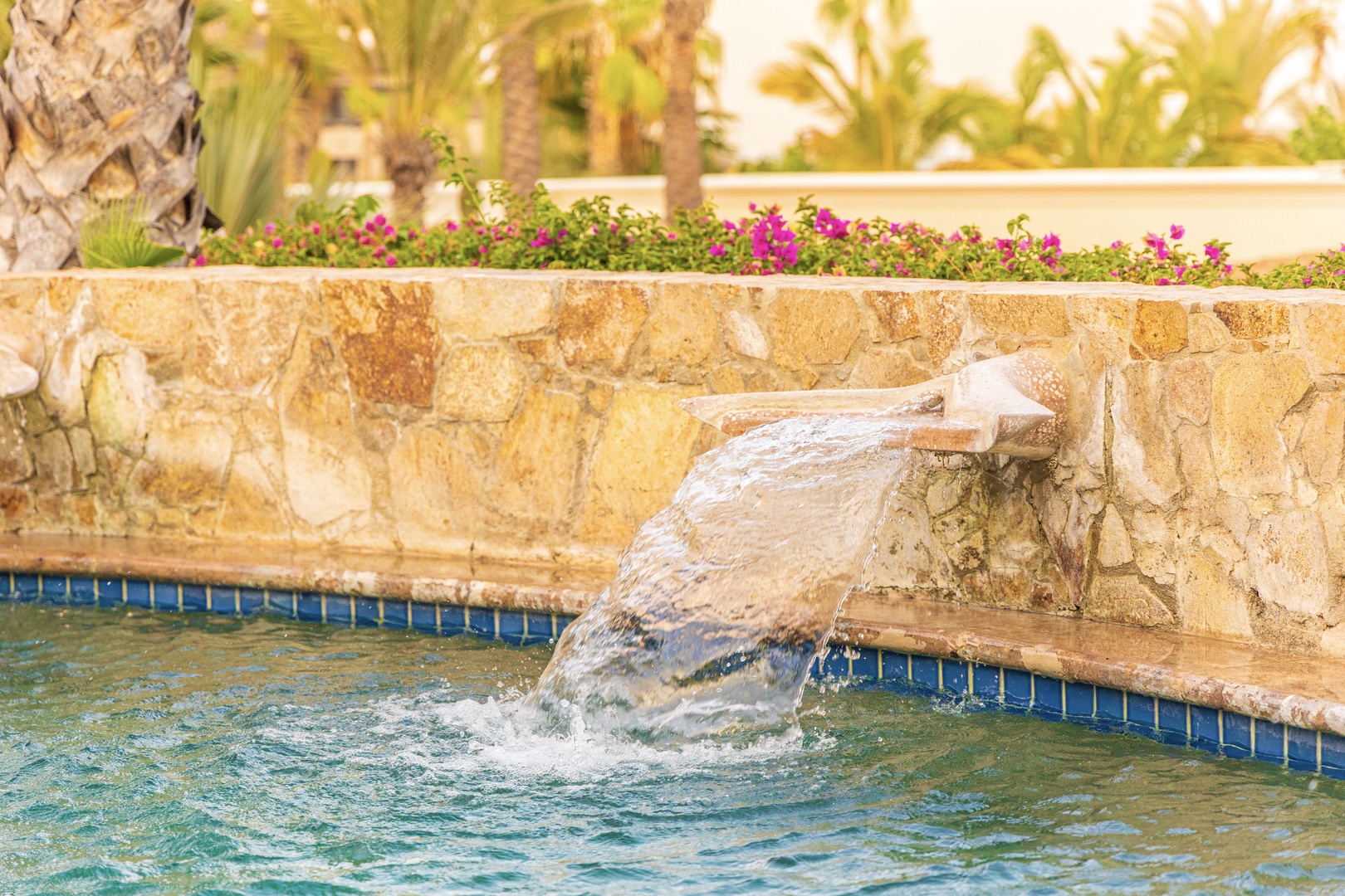 A cascading water feature adds a soothing soundtrack to the tropical pool area, surrounded by colorful blooms and swaying palms—a perfect spot to relax and unwind.