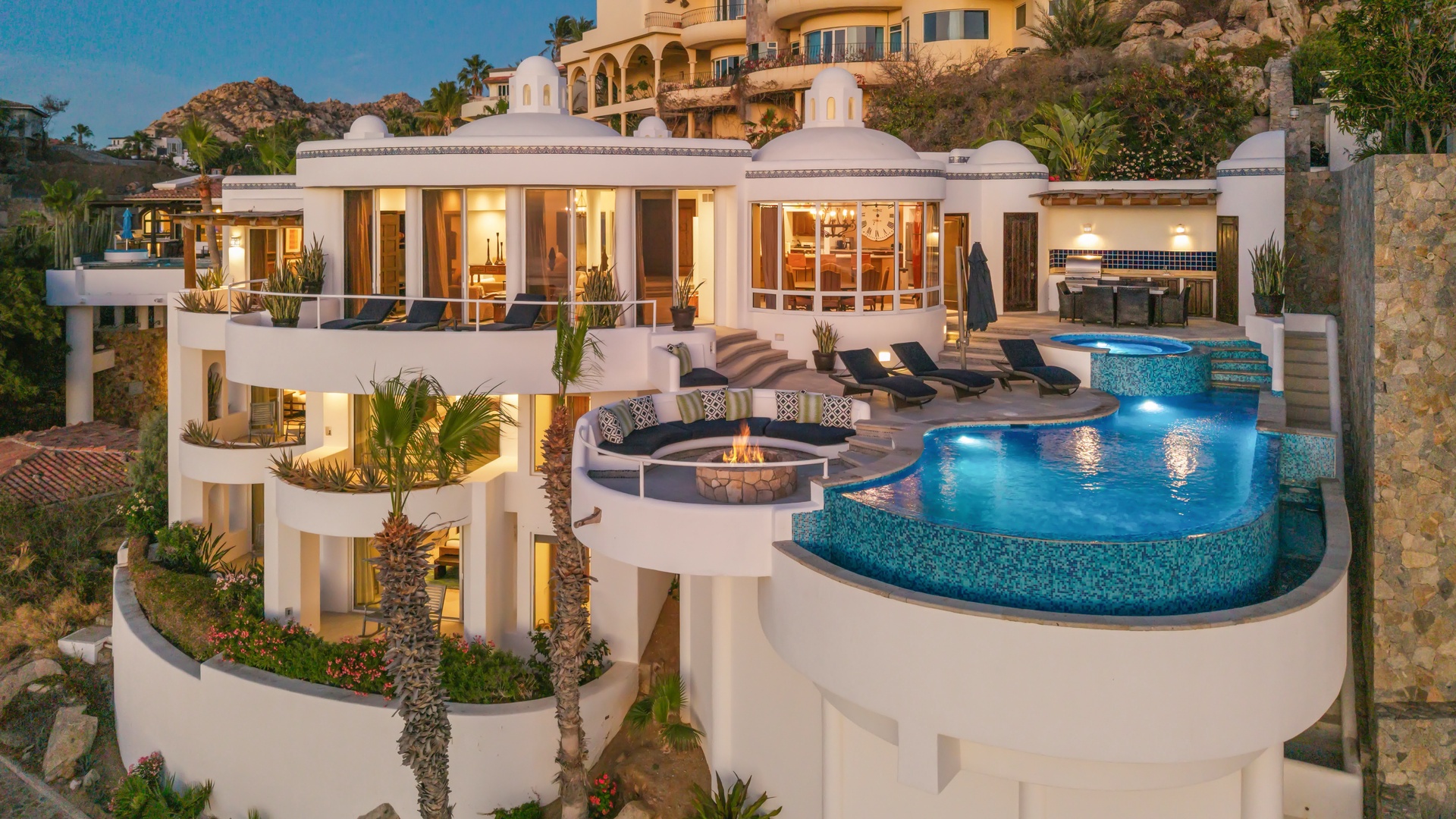 Stunning luxury villa featuring multiple terraced levels, infinity pool with water feature, and outdoor fire pit seating area surrounded by tropical landscaping.