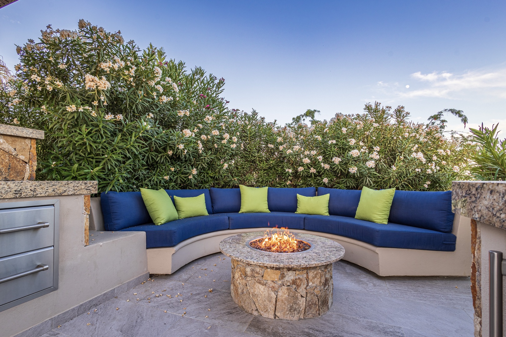 Unwind in this serene outdoor retreat, where a cozy curved sectional wraps around a crackling fire pit, surrounded by lush greenery and open sky—your perfect evening escape.