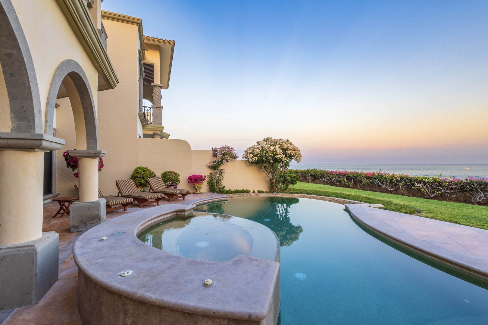 Mediterranean-style villa with curved pool, lush landscaping, and beachfront access captured during the golden hour of early evening.