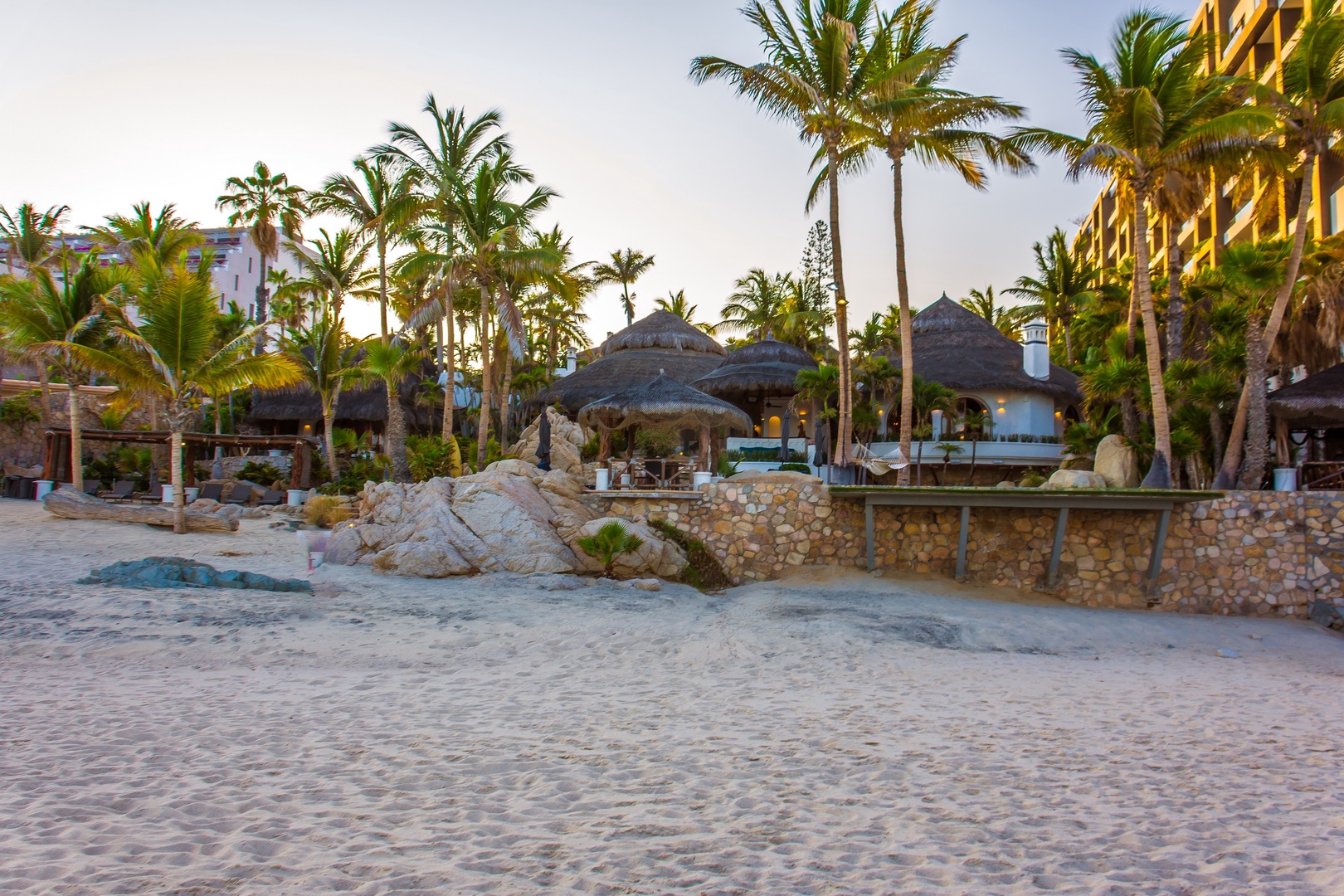 Beachfront resort property nestled among swaying palms with sandy shore access and thatched-roof dining pavilions nearby, capturing the golden hour glow in a tropical coastal setting.