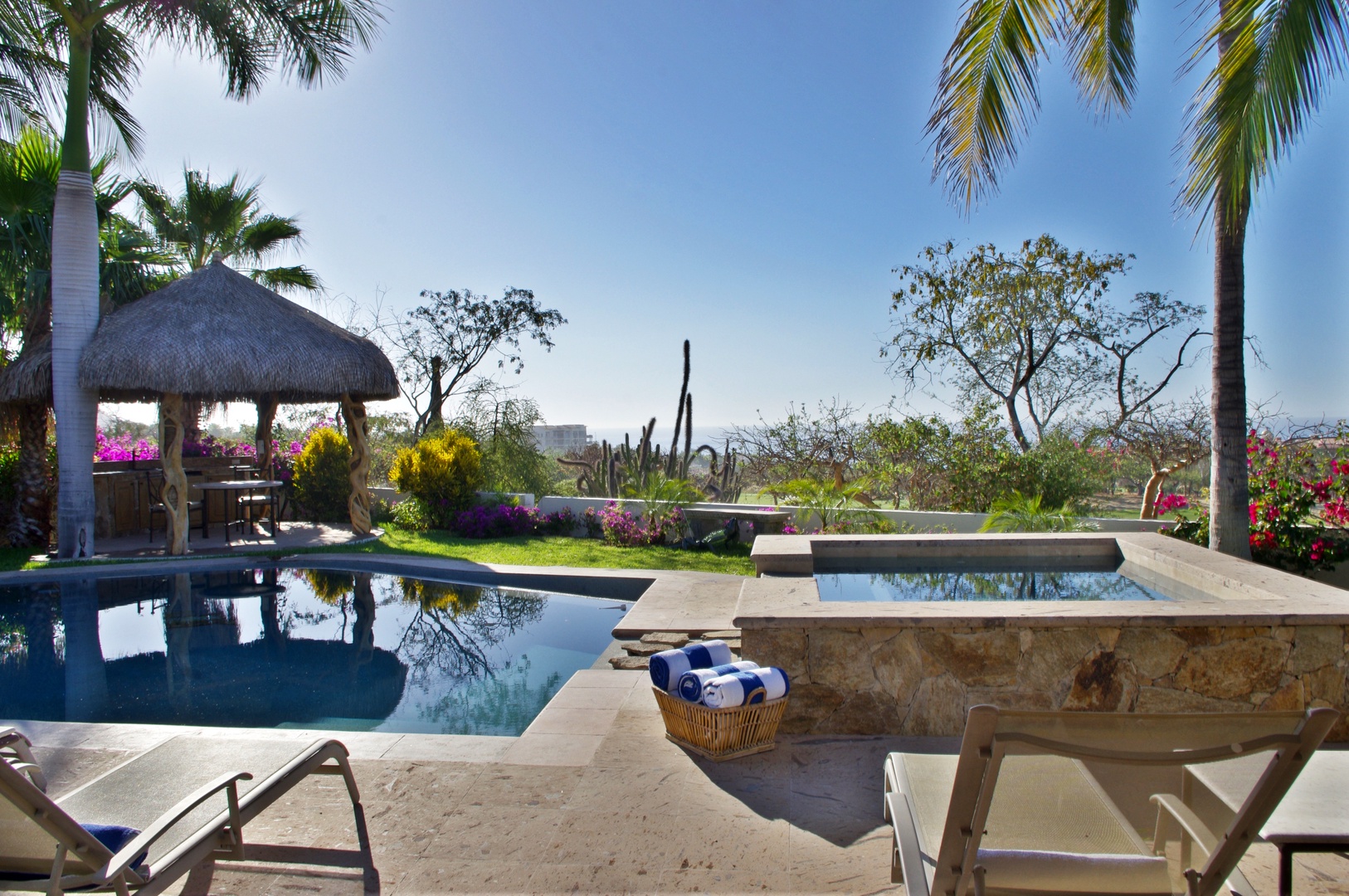 A tropical paradise where two pools await—one for leisurely swims, the other a raised hot tub. Lounge on sunbeds under swaying palms, surrounded by vibrant bougainvillea and open views.
