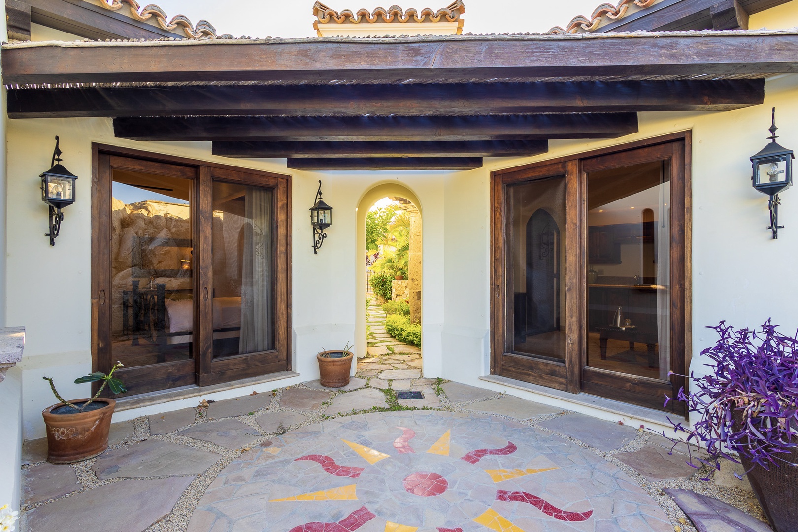 Mediterranean-style entrance courtyard featuring exposed wood beam pergola, terracotta planters with greenery, decorative mosaic tile flooring, and an arched garden passageway offering glimpses of lush landscaping beyond.