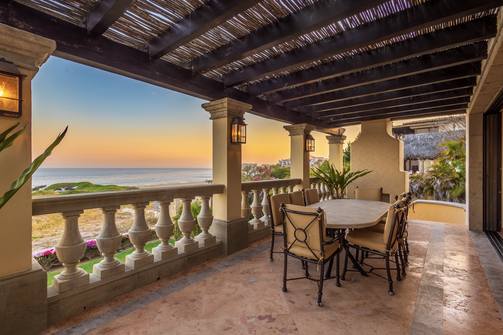 Sunset unfolds in warm amber hues over the ocean from this covered terrace, where your outdoor dining space invites evening meals beneath natural wood beams and glowing lanterns.