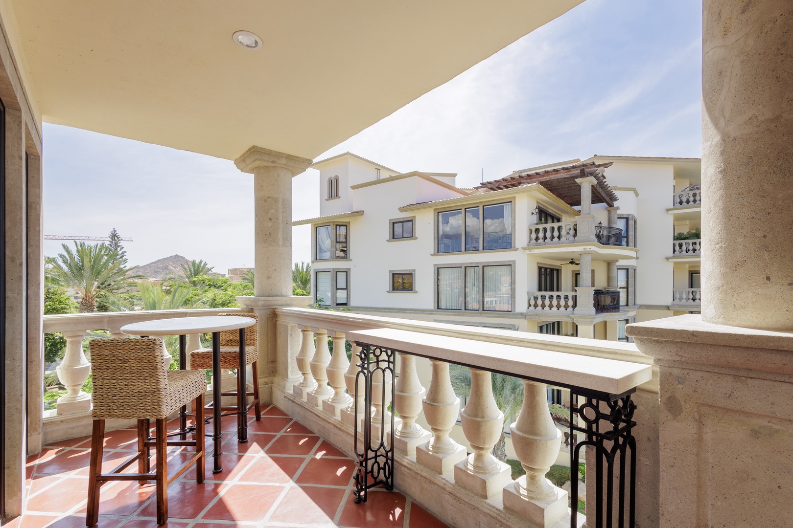 Elegant covered terrace with bistro seating overlooking tropical palms and mountain views. Red-tiled flooring and decorative columns frame a tranquil scene perfect for morning coffee or evening cocktails.