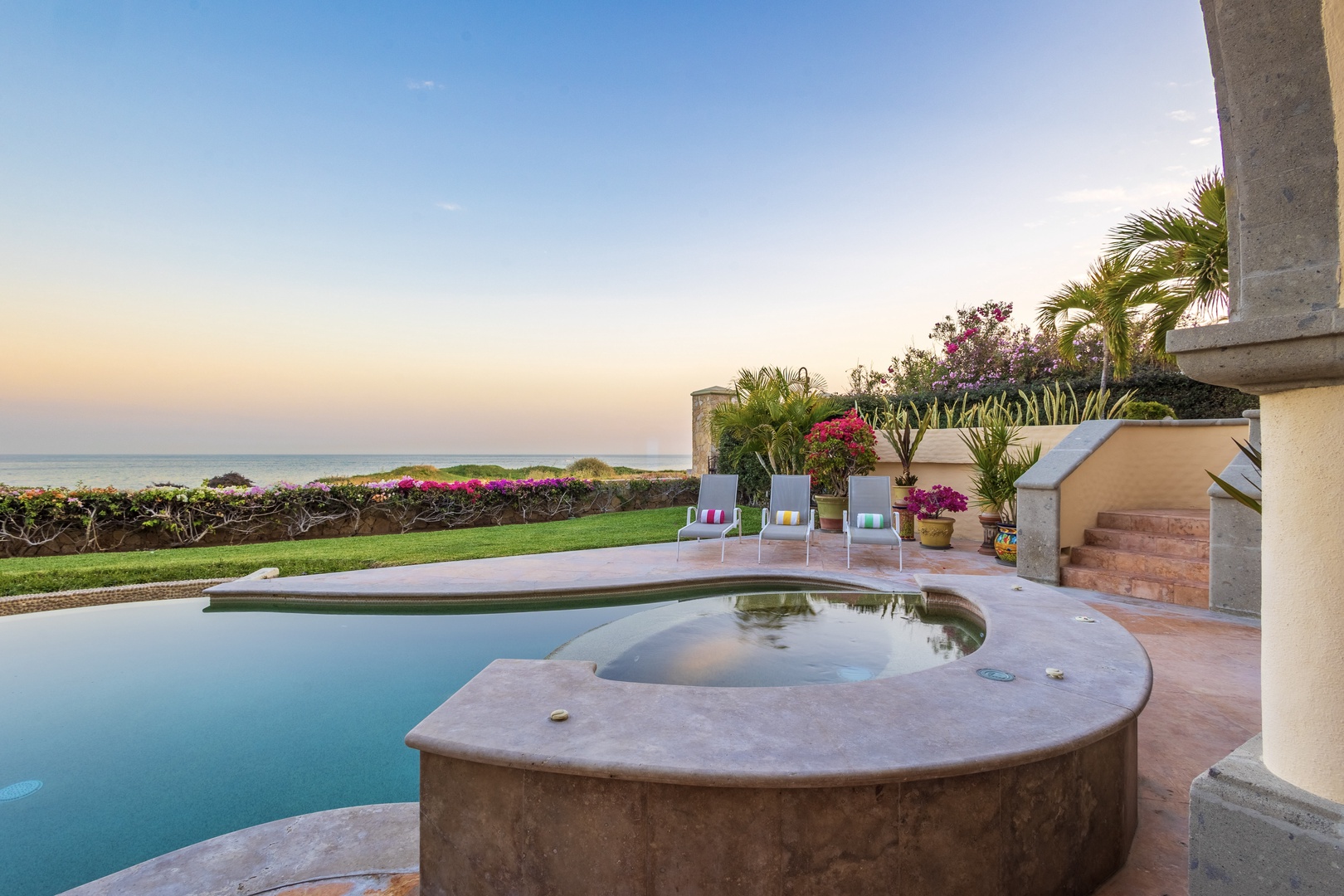 Ocean-view paradise with a sparkling pool, hot tub, and lush gardens. The property showcases stunning sea vistas and manicured lawns under a pastel sunset sky.