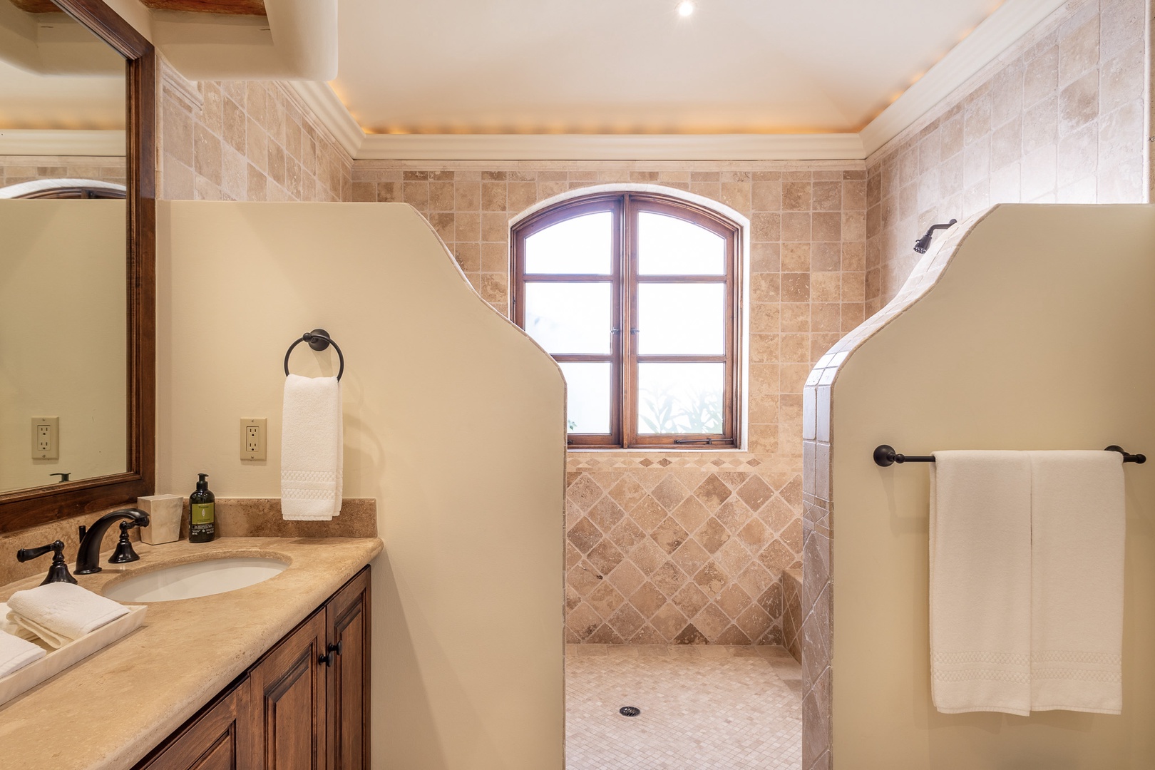 Step into your private spa-like retreat with a spacious walk-in shower surrounded by beautiful travertine tile, where natural light pours through an arched window, making each morning feel serene and refreshing.