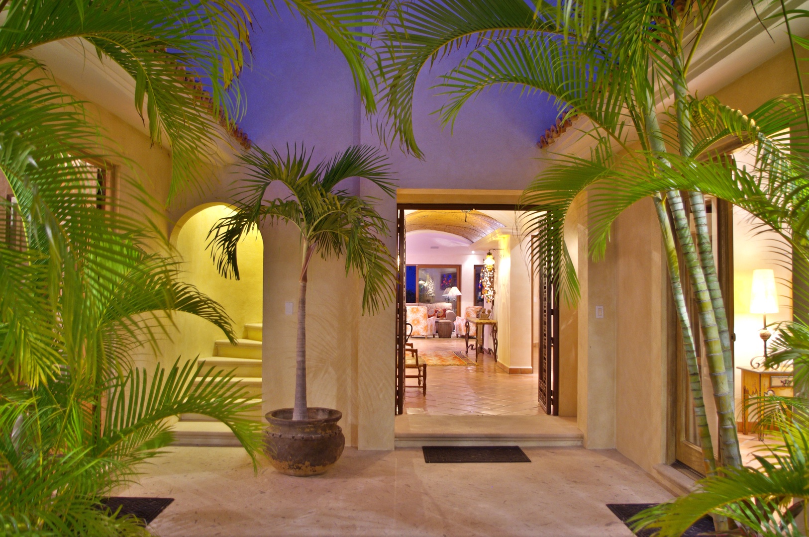 A welcoming entrance framed by lush tropical palms opens into a warmly lit interior, blending indoor comfort with outdoor elegance as twilight settles over the property.