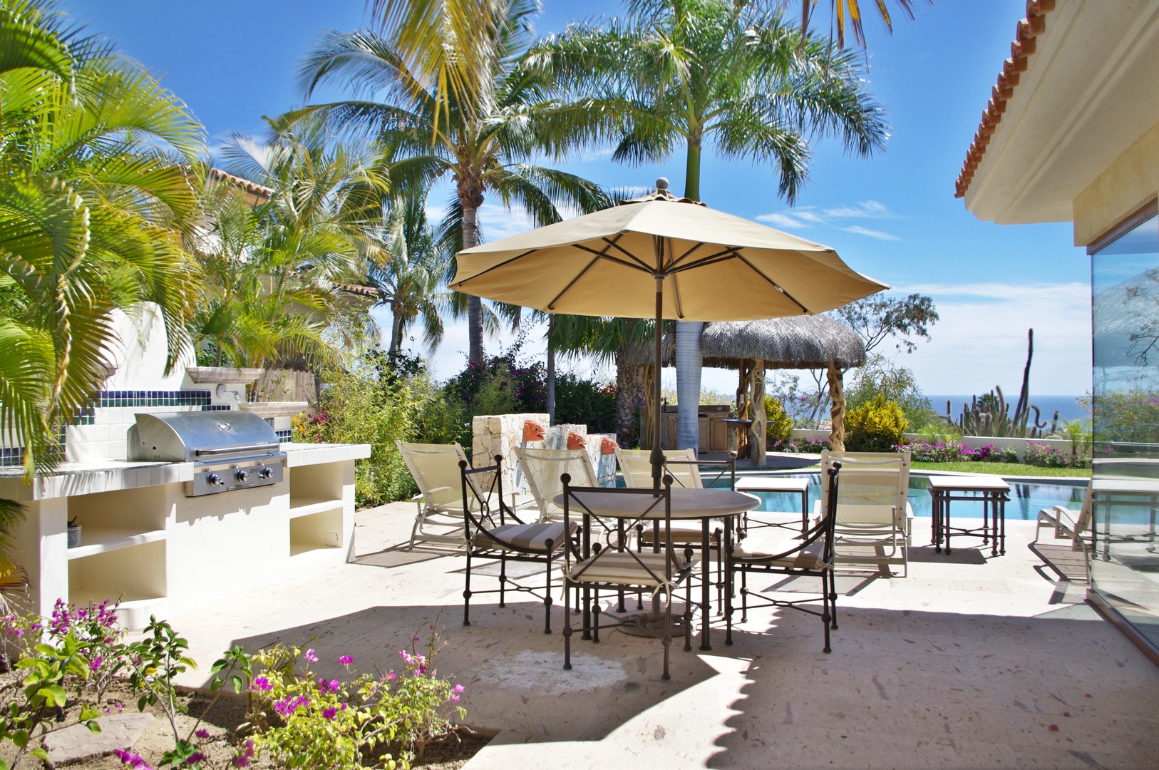 A sun-drenched patio features a spacious dining area under a large umbrella, with a built-in grill and sparkling pool nearby. Tropical palms and vibrant flowers frame your outdoor retreat, perfect for meals and relaxation.