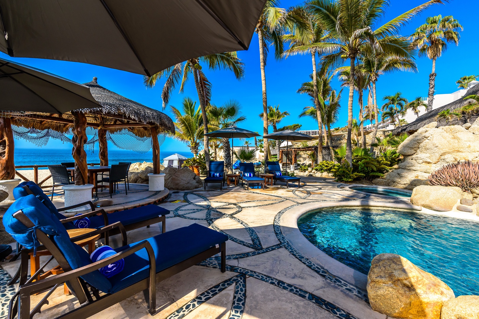 A beachfront resort property featuring tropical landscaping, thatched palapas, and a sparkling pool surrounded by natural stone and lush palm trees under clear blue skies.