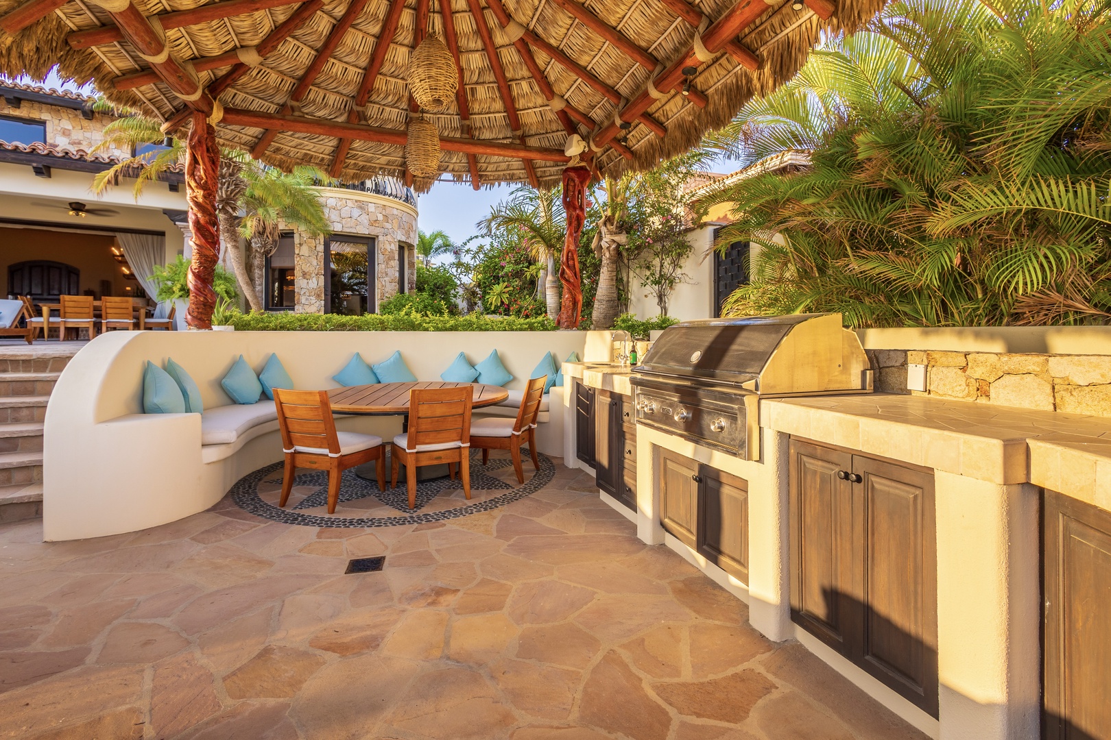 Your own tropical outdoor kitchen awaits under a palapa, with built-in grill and curved seating perfect for sun-soaked meals surrounded by lush palms and garden views.