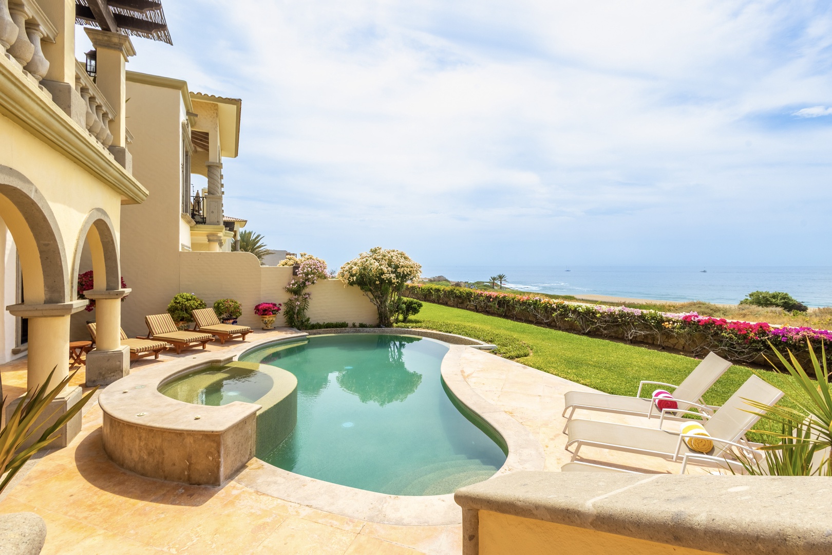 Beachfront villa with private pool, spa, manicured lawn, and panoramic ocean views. Mediterranean-style architecture with palm trees and colorful gardens frames the coastal setting.