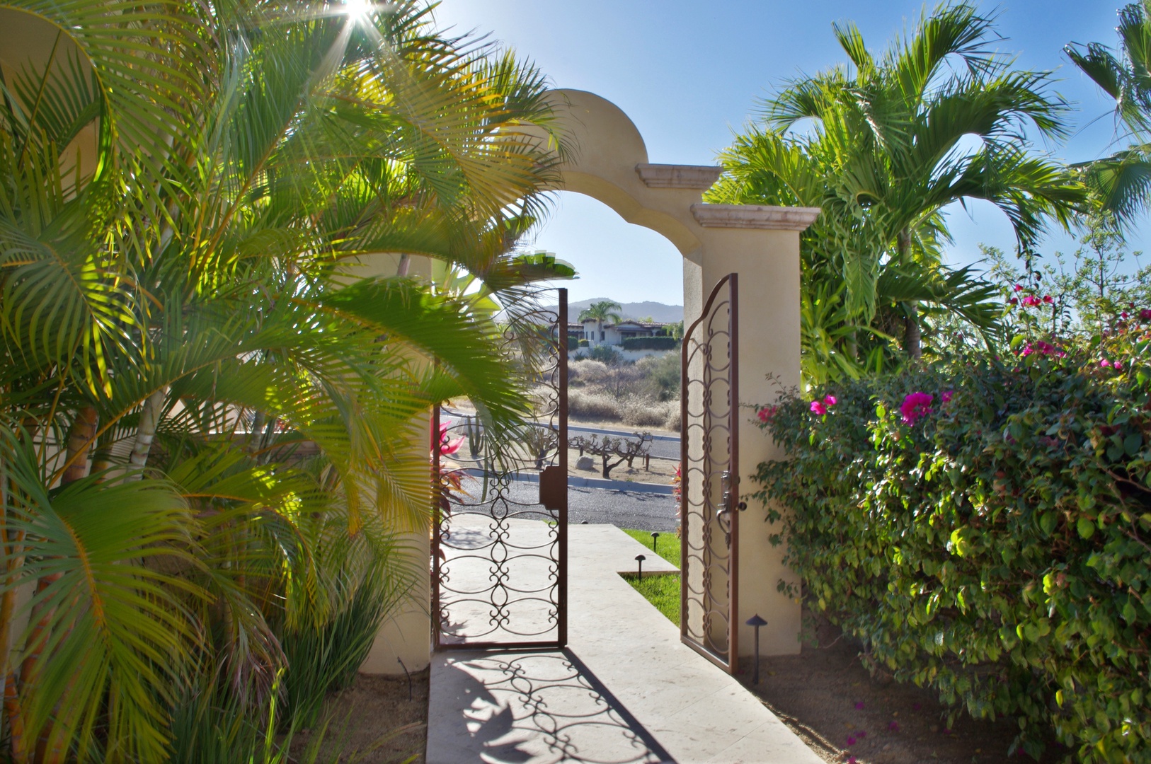 Elegant entry gate framed by lush palm trees and vibrant bougainvillea welcomes guests to this desert oasis, where tropical beauty meets sunny serenity.