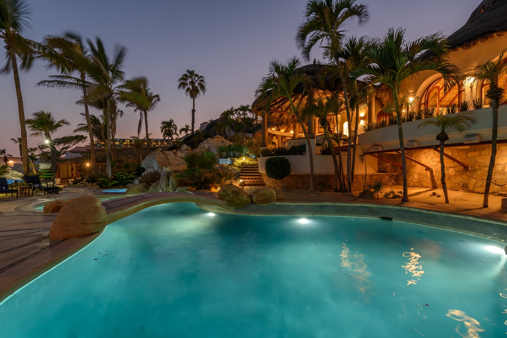 A luxurious tropical resort features a stunning illuminated pool surrounded by lush palms and natural rock formations, set against a tranquil evening sky.