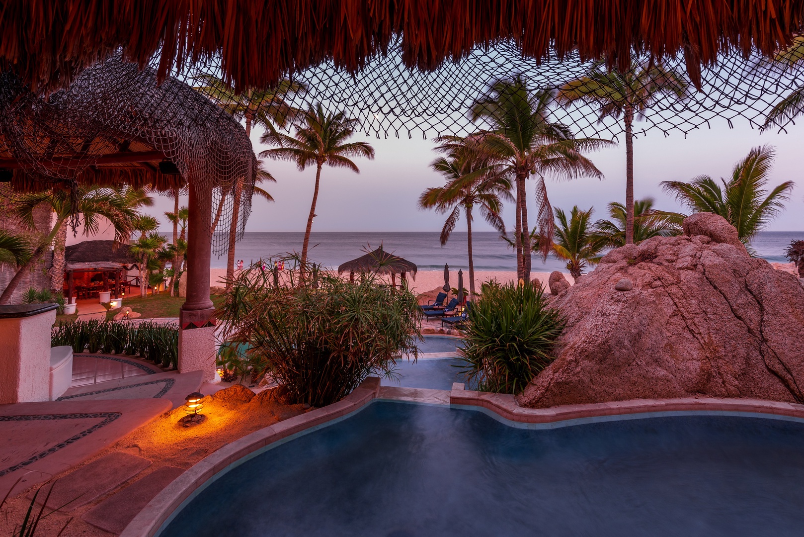 A breathtaking beachfront oasis features a stunning pool framed by swaying palms and thatched palapas, perfectly positioned where tropical paradise meets the sea at dusk.