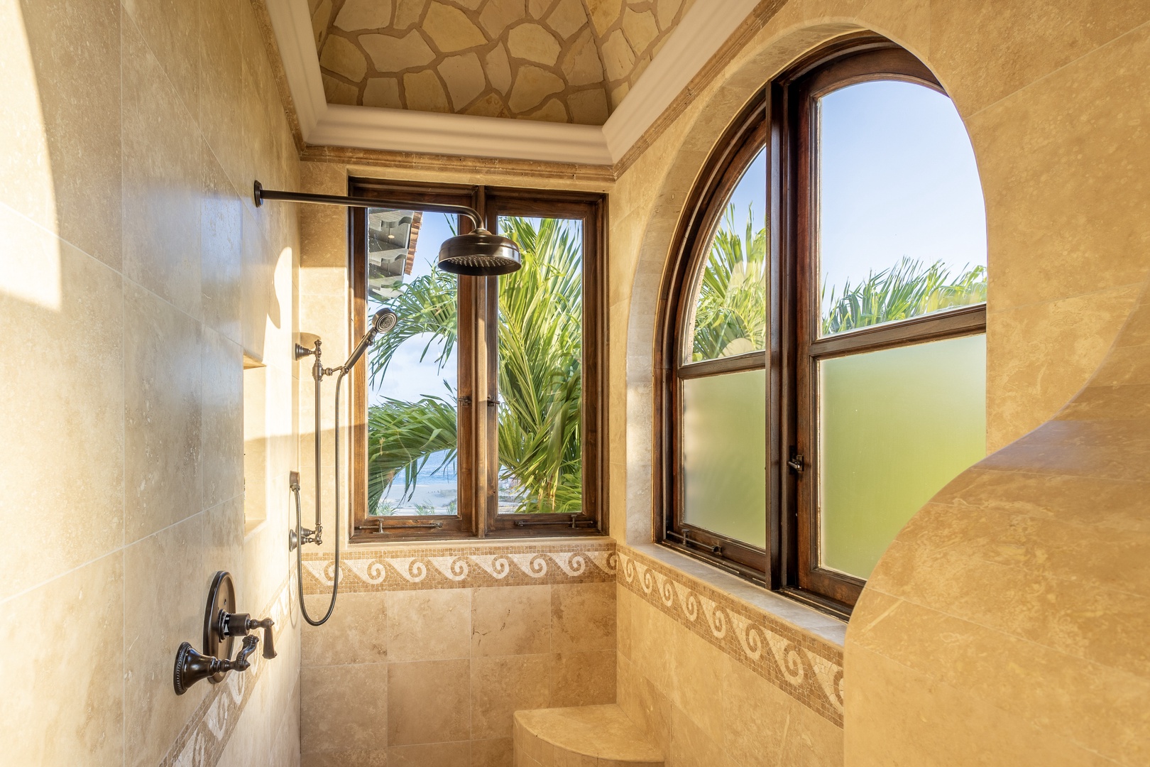 Step into your private spa-like shower with elegant travertine walls, a decorative ceiling, and an arched window framing tropical palms and glimpses of the ocean beyond.