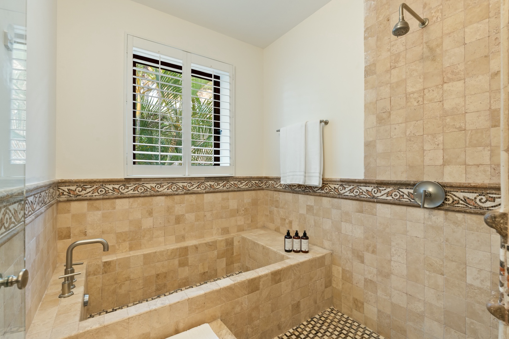Unwind in your luxurious spa-style bathroom featuring elegant travertine finishes, a deep soaking tub, and tropical garden views through the window.