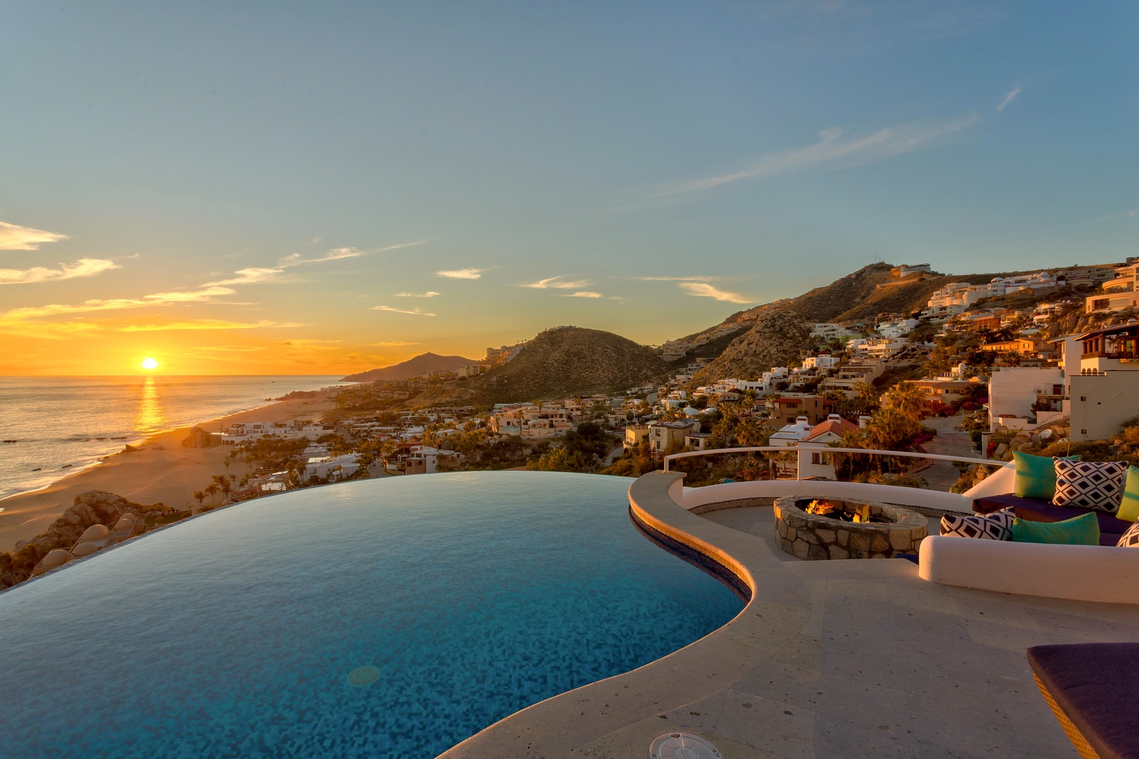 This coastal property overlooks a hillside community and the Pacific Ocean, with an infinity pool, outdoor seating, and fire pit. The setting captures golden hour light across the water and mountains.