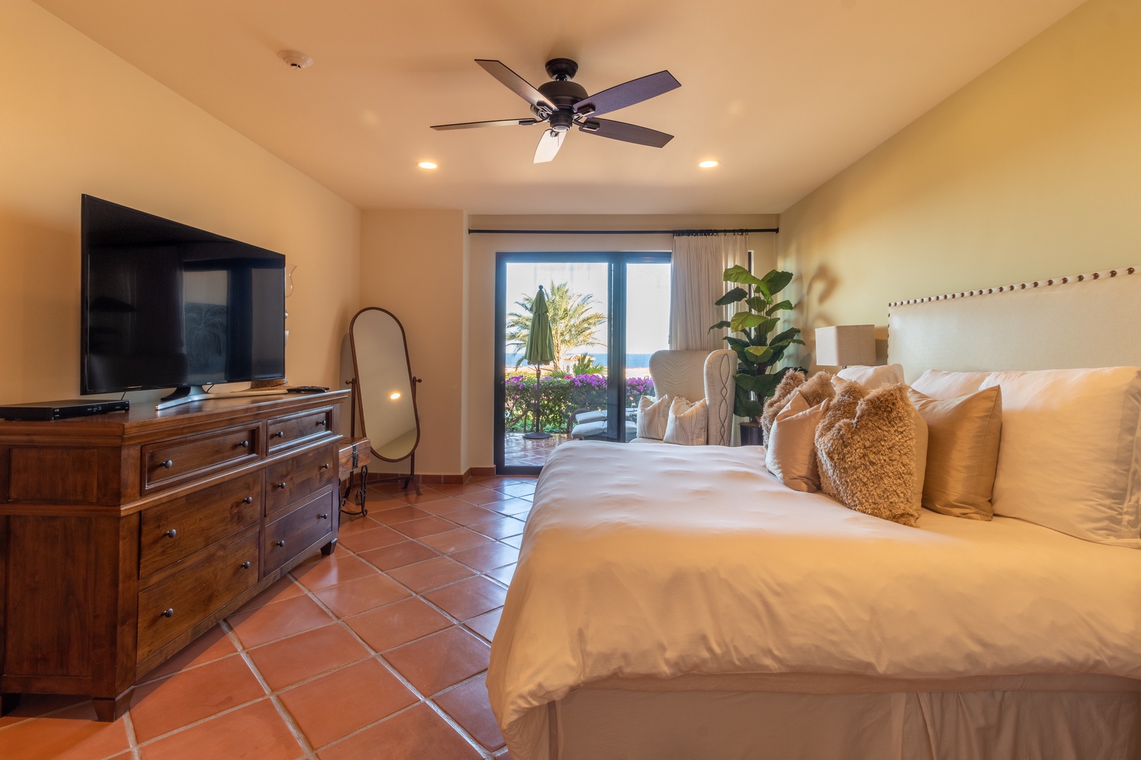 Your airy bedroom retreat features plush linens, terracotta tile floors, and a private doorway framing vibrant gardens and ocean views—perfect for unwinding in coastal comfort.
