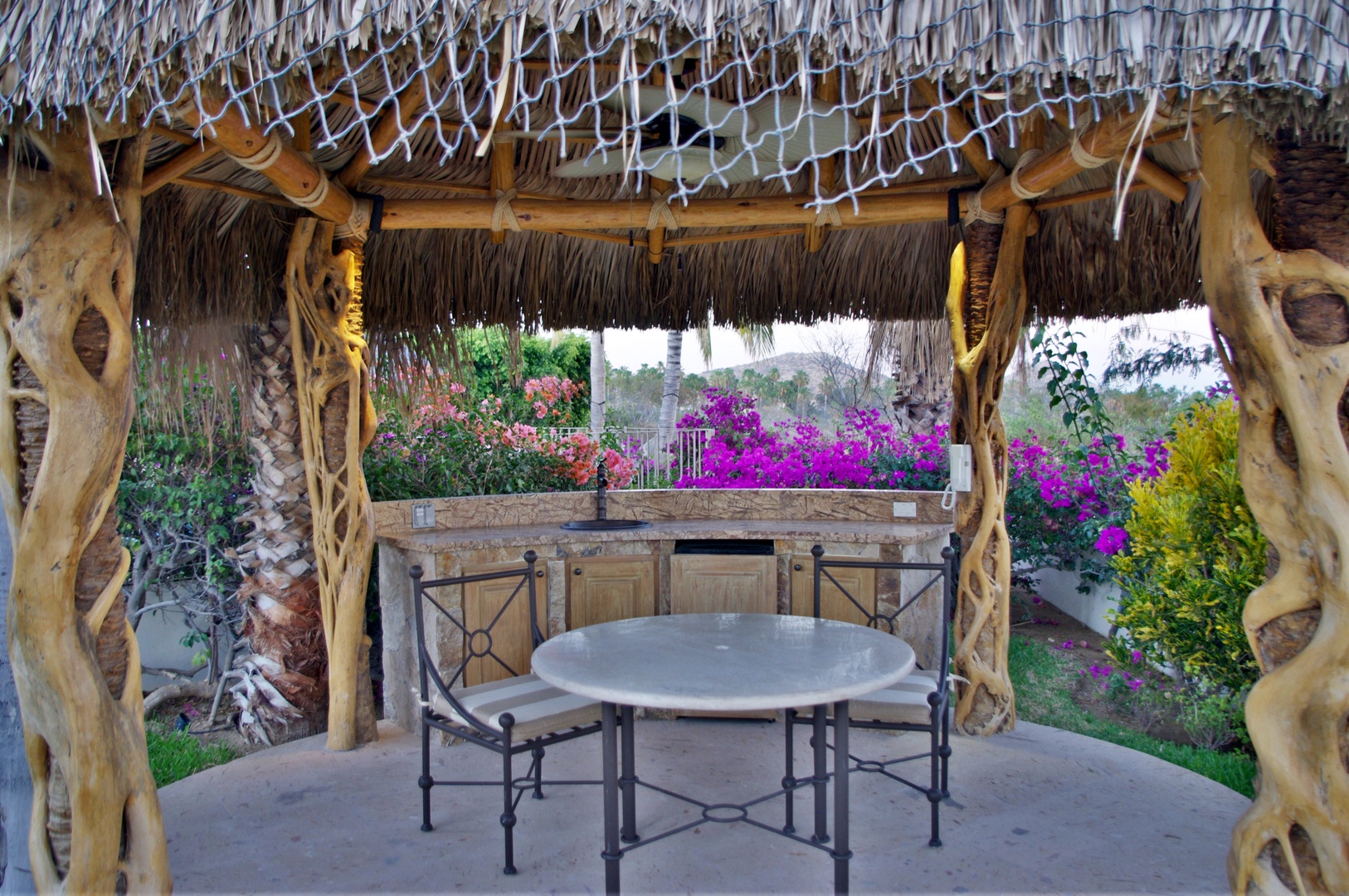A tropical palapa with rustic wood pillars frames your outdoor dining space, where vibrant bougainvillea blooms create a colorful backdrop for alfresco meals in the garden.