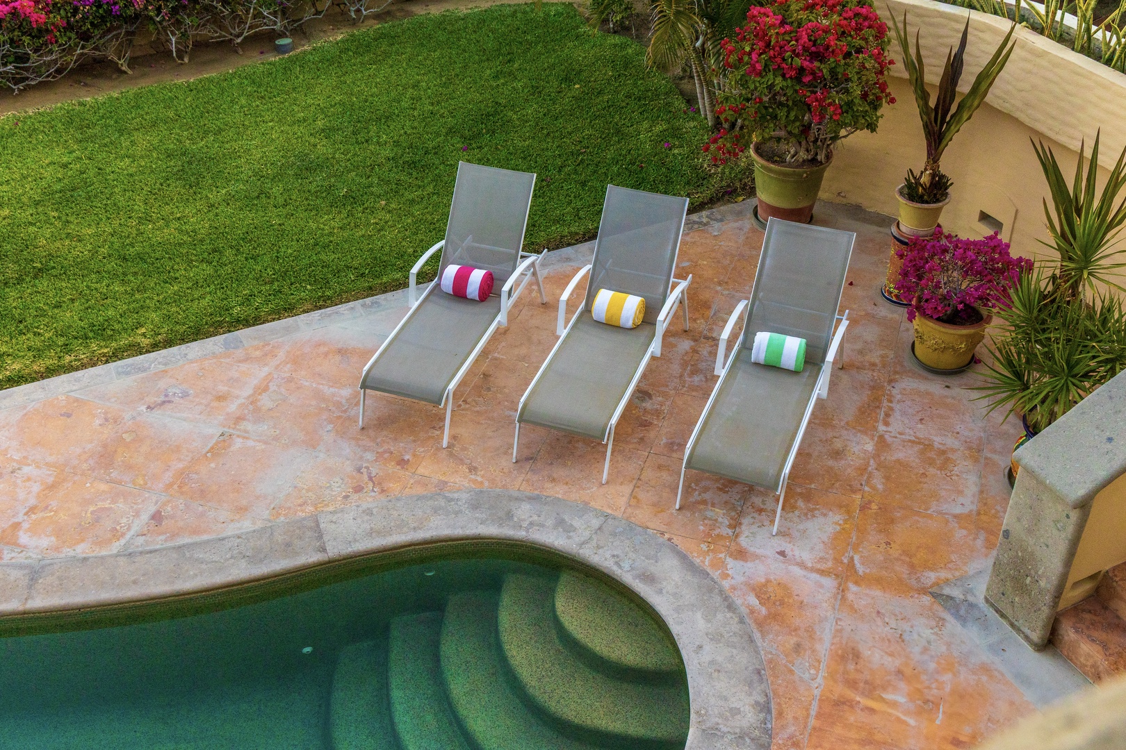 The property features a sparkling pool surrounded by lush lawn, vibrant gardens, and comfortable loungers for sun-soaked relaxation in a tropical setting.