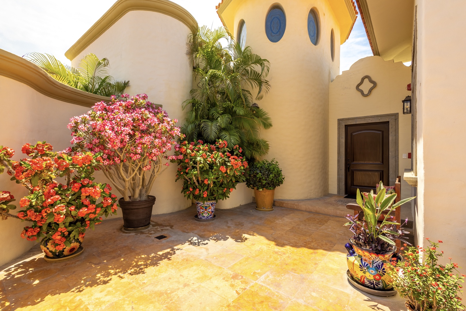 Mediterranean-style villa entrance featuring a tower with circular windows, vibrant bougainvillea, tropical palms, and colorful decorative planters. The private courtyard welcomes guests with Spanish Colonial architectural details and lush landscaping.
