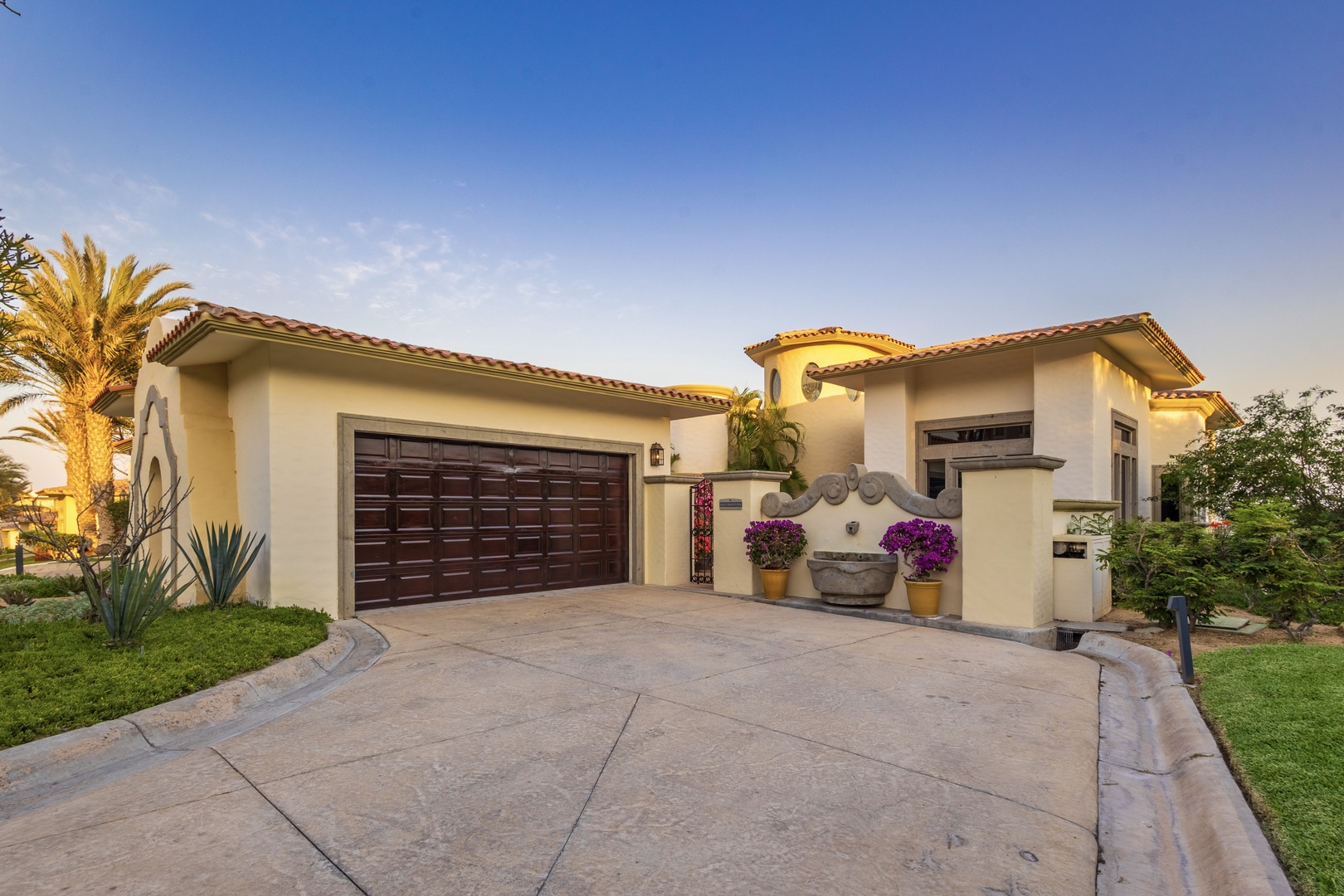 Mediterranean-style villa with terra-cotta tile roof, vibrant purple florals, tropical palms, and manicured landscaping, photographed during golden hour.
