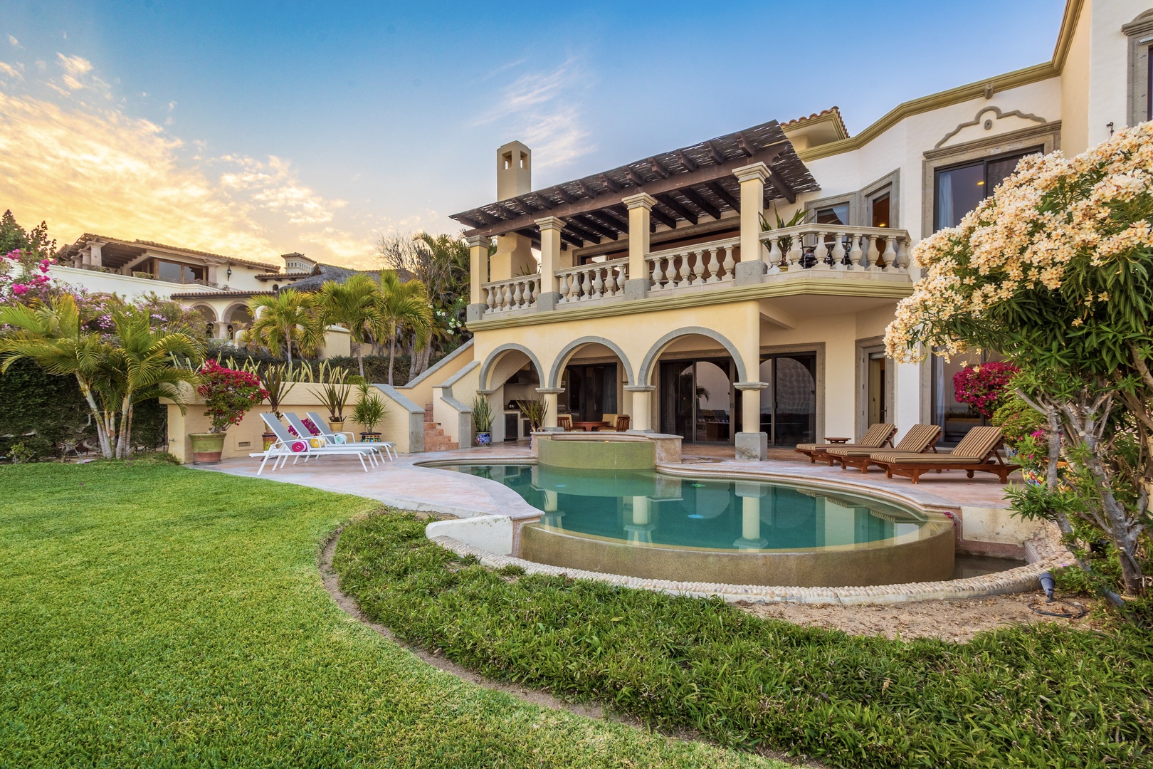 An elegant Spanish-style villa showcasing architectural beauty with arched columns, lush tropical gardens, and private pool area, all set beneath a dramatic evening sky in an exclusive resort community.