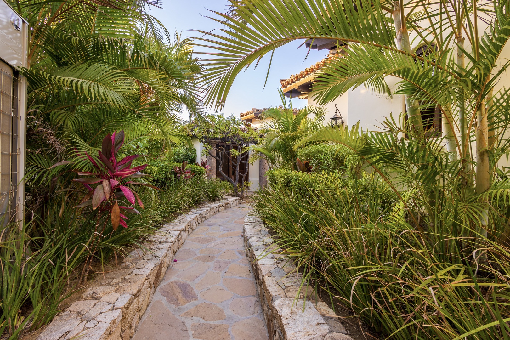 A sun-drenched stone pathway winds through lush tropical gardens, welcoming you to your private retreat beneath swaying palms and vibrant foliage.