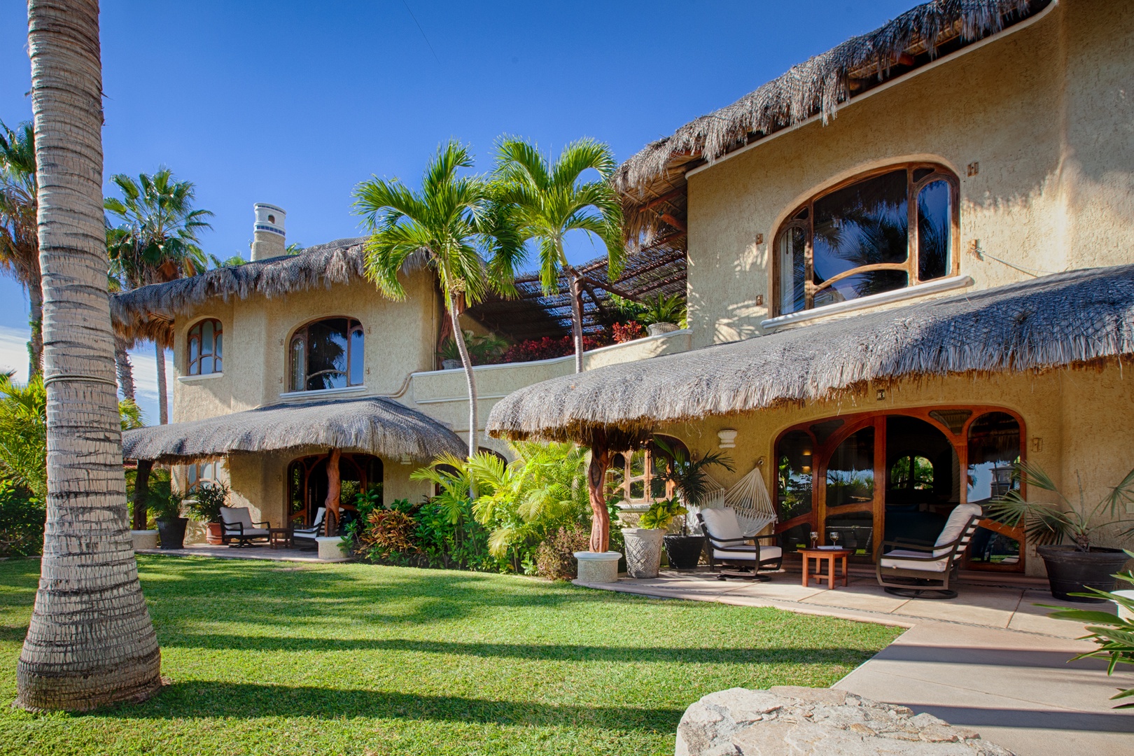 Tropical paradise villa with thatched roof architecture, surrounded by palm trees and lush gardens under blue skies.