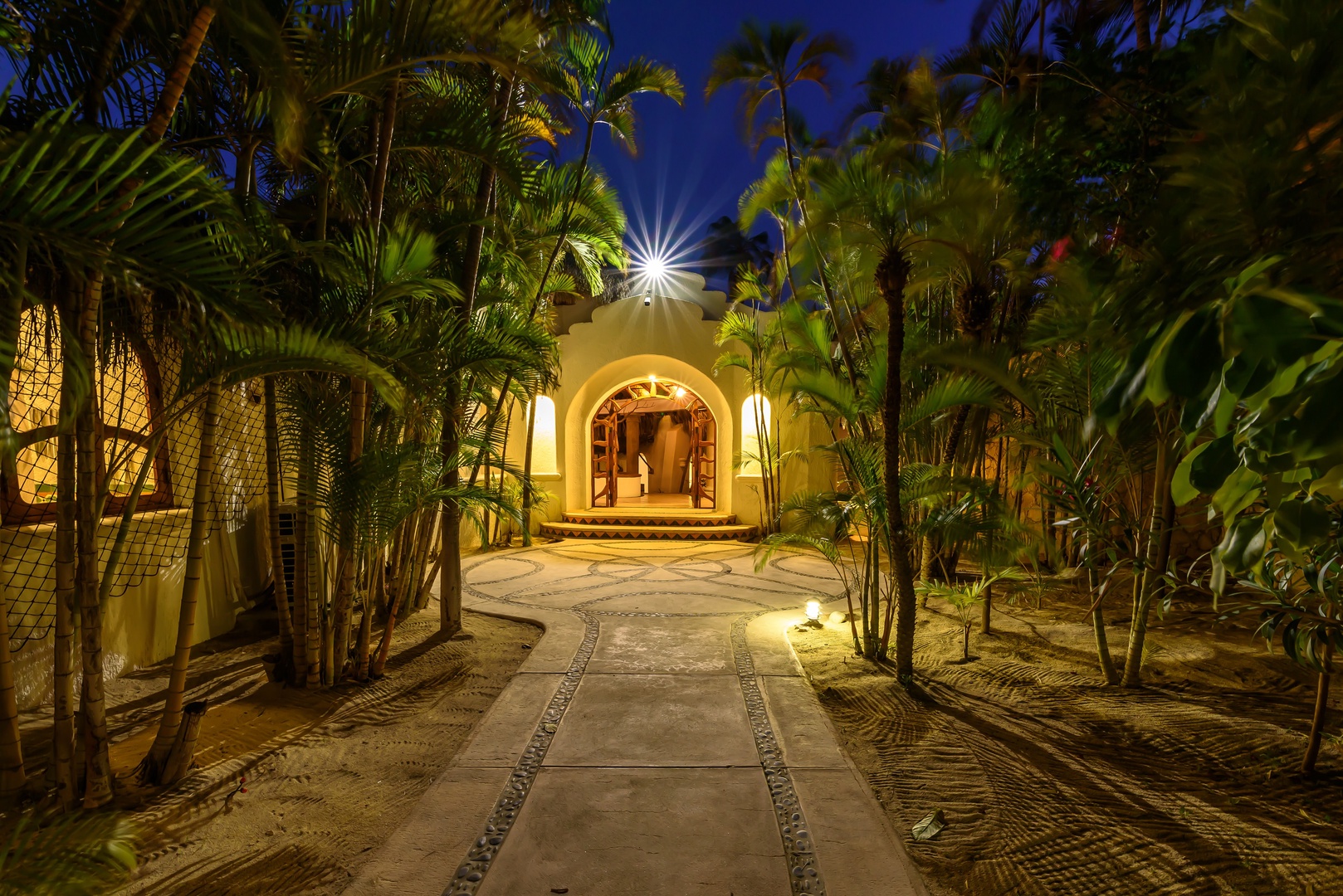 A palm-lined pathway leads to an illuminated arched entrance, creating an enchanting tropical arrival experience under twilight skies.