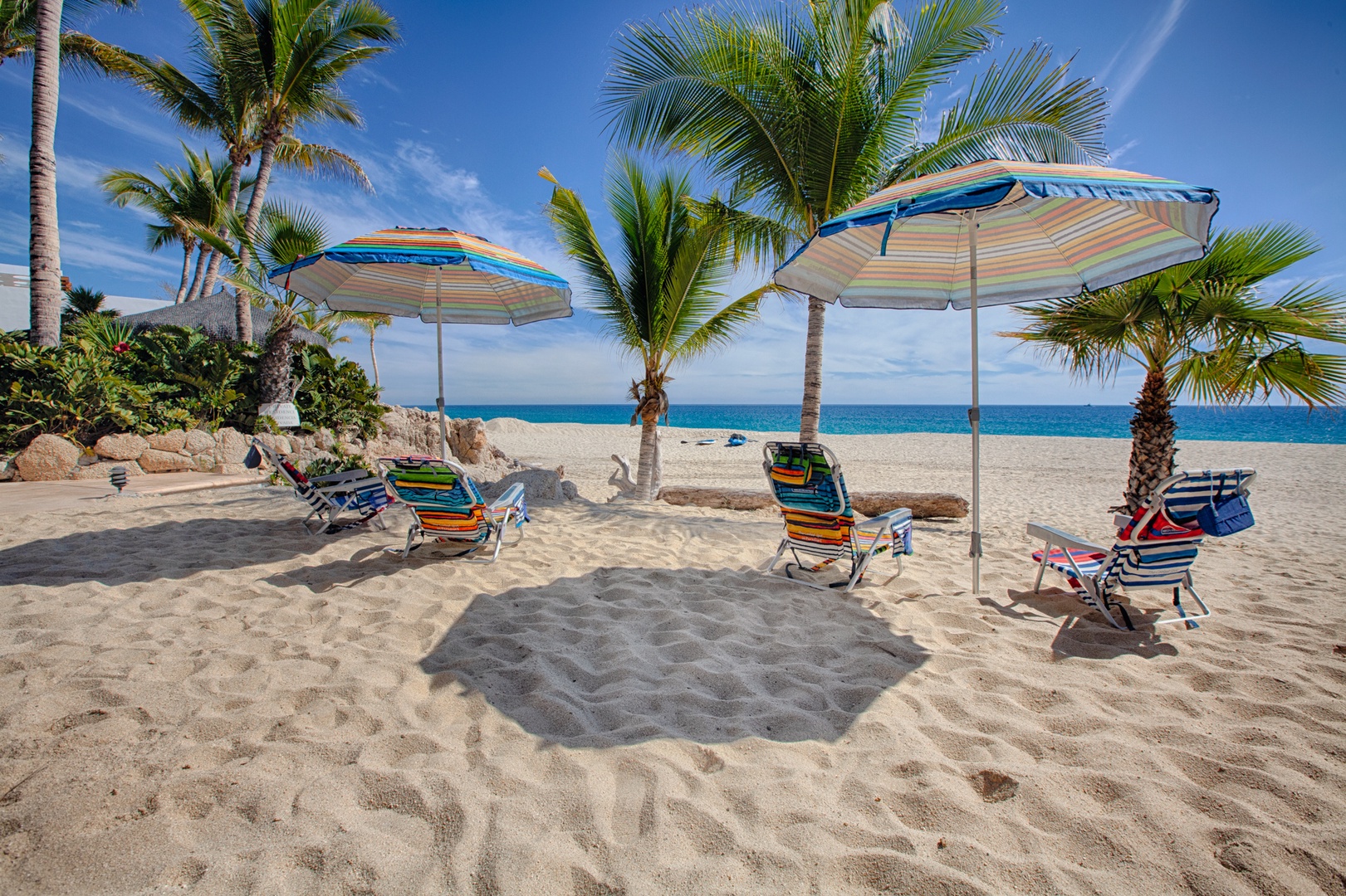 Your personal beach setup awaits with colorful umbrellas and comfy chairs on pristine sand, framed by swaying palms and turquoise waters.