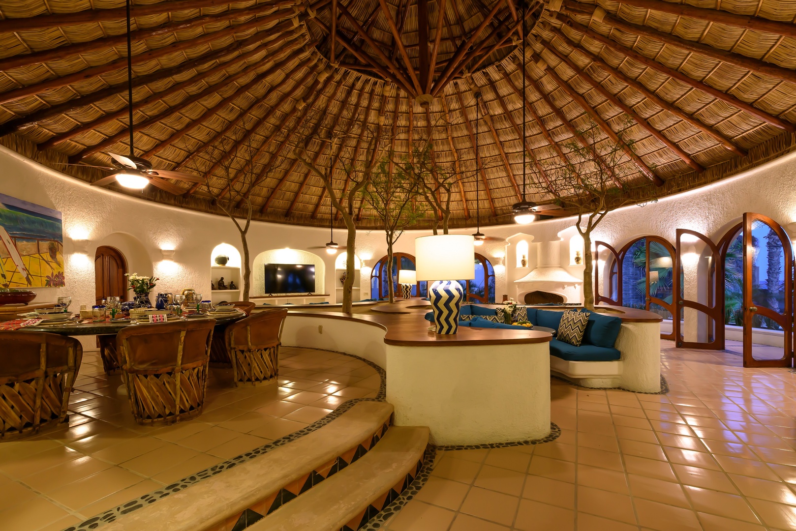 Step into this stunning palapa-style living space where natural textures meet modern comfort. Lounge on curved seating under a beautifully crafted thatched roof, gather around the dining table, and feel the tropical evening breeze through open archways.
