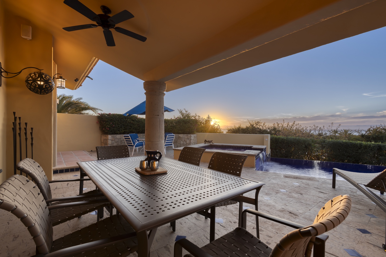 Your private outdoor dining retreat awaits under a covered patio with ceiling fan, featuring an elegant table set against stunning sunset views over lush gardens.