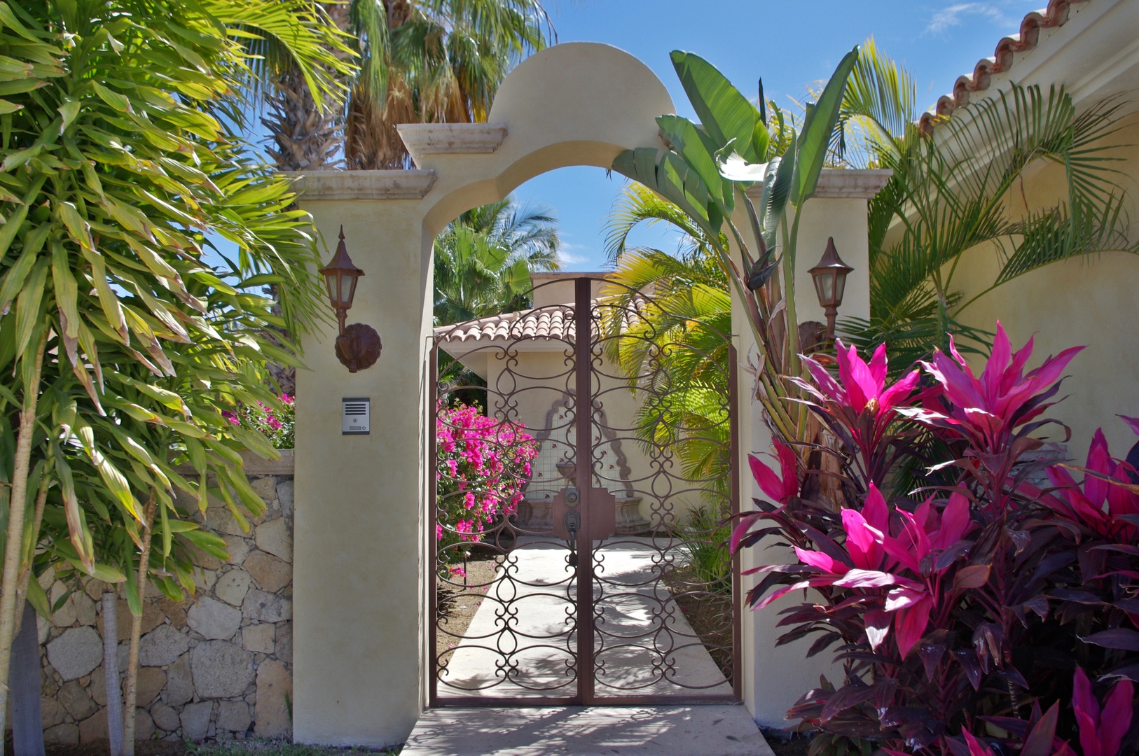 This charming Mediterranean-style entrance is framed by lush palms, vibrant bougainvillea, and striking tropical plants, welcoming guests to a sun-drenched coastal retreat.