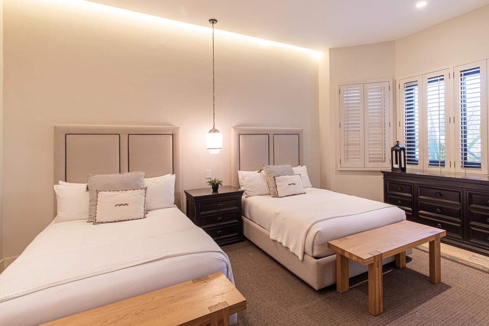 Your serene bedroom awaits with twin beds dressed in plush white linens, soft ambient lighting creating a warm glow, and natural light filtering through elegant shutters for your perfect restful retreat.