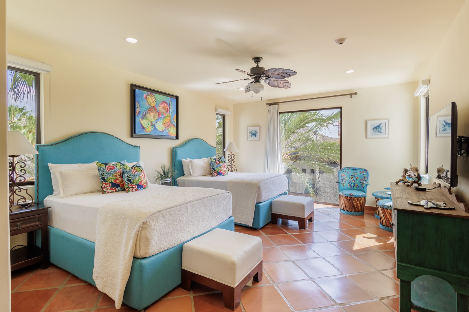 Bright, coastal-inspired room featuring twin beds with turquoise headboards, tropical ceiling fan, and sunny windows framing palm views—your serene retreat awaits.