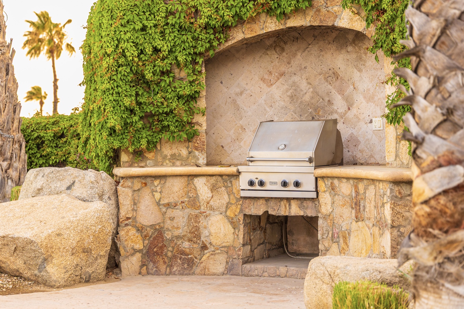 A built-in grill station awaits your outdoor cooking adventures, framed by lush greenery and palm trees under the warm desert sun—perfect for grilling your favorite meals in style.