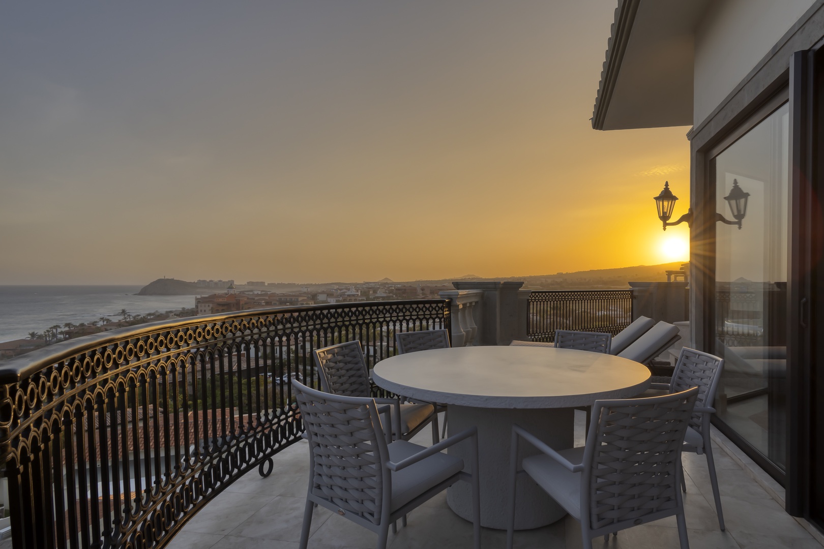 Sunset view from balcony overlooking the coastline and distant islands, featuring outdoor dining area with decorative ironwork railing and warm ambient lighting.
