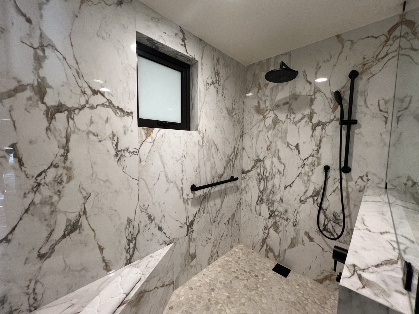 Stunning marble-clad shower with rainfall and handheld heads, featuring elegant black fixtures and a textured pebble floor—your personal spa retreat.