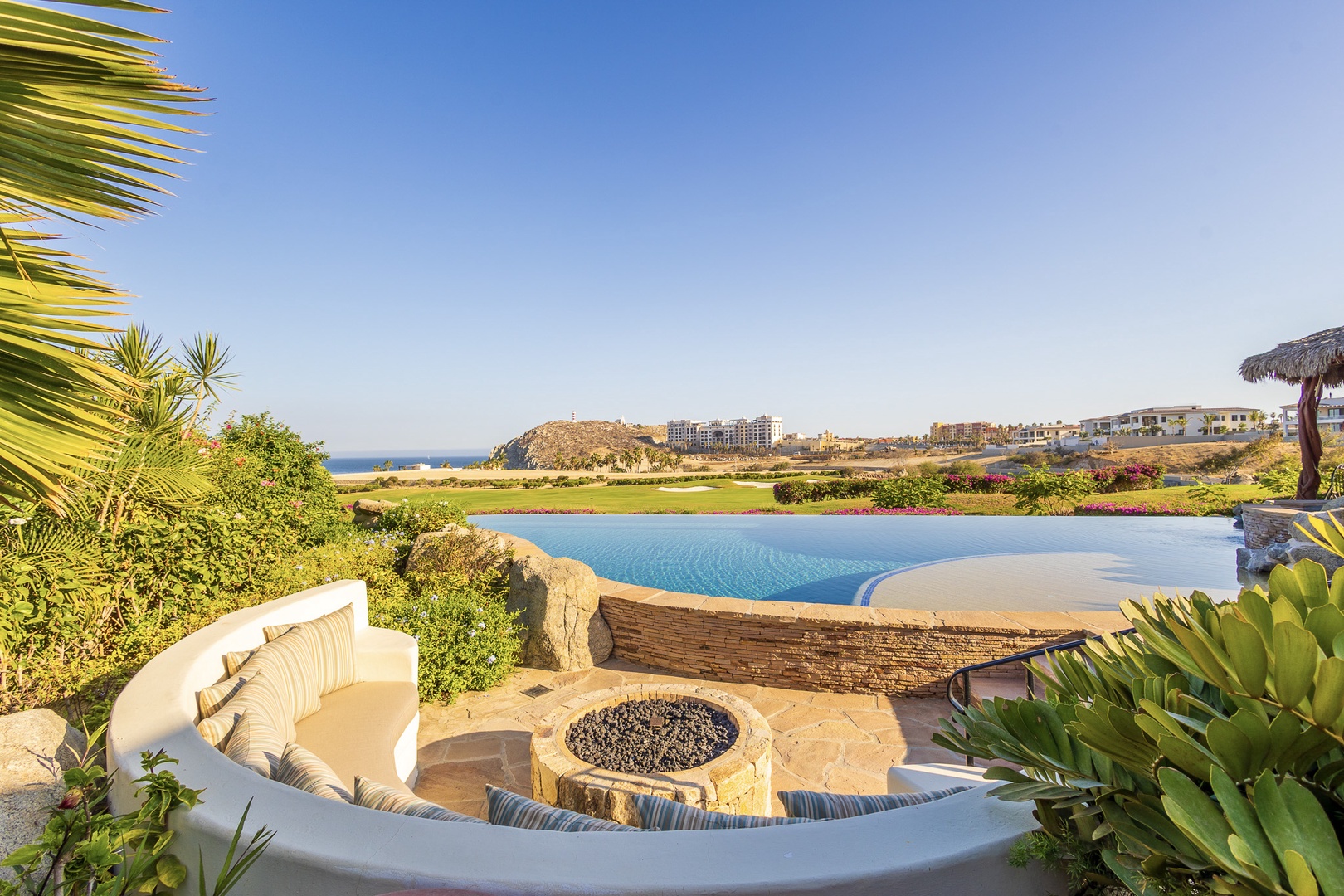 Curved seating wraps around a stone fire pit, overlooking an infinity pool with breathtaking golf course and ocean views—your perfect spot to unwind.