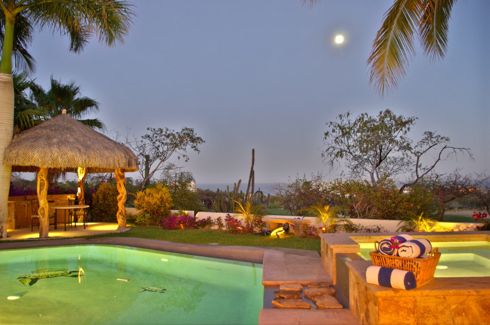 The property features a beautifully illuminated pool area surrounded by lush tropical landscaping, creating a serene evening ambiance beneath the moonlit sky.