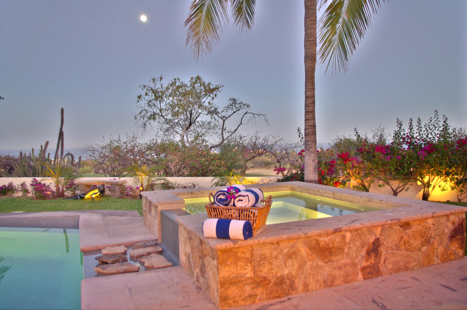 Serene desert oasis features illuminated hot tub surrounded by lush gardens, palm trees, and vibrant bougainvillea under moonlit sky—a perfect evening escape.