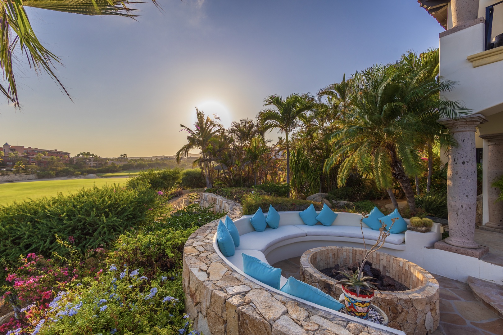 Luxurious outdoor terrace overlooking manicured golf course with curved seating, stone fireplace, lush palm trees, and vibrant garden at golden hour. Part of an upscale resort property with striking architectural columns.